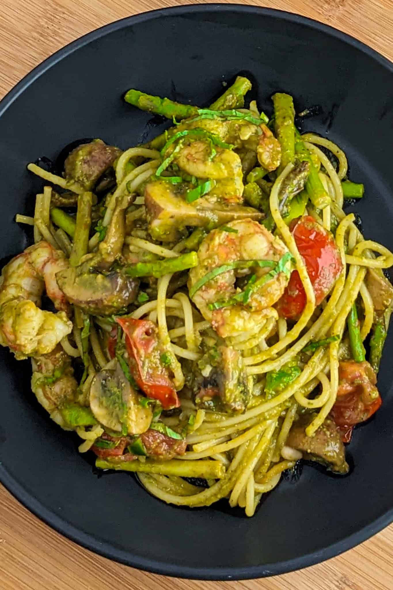 Vibrant Argentinian shrimp pesto pasta with asparagus, mushrooms, and cherry tomatoes on a dark plate.